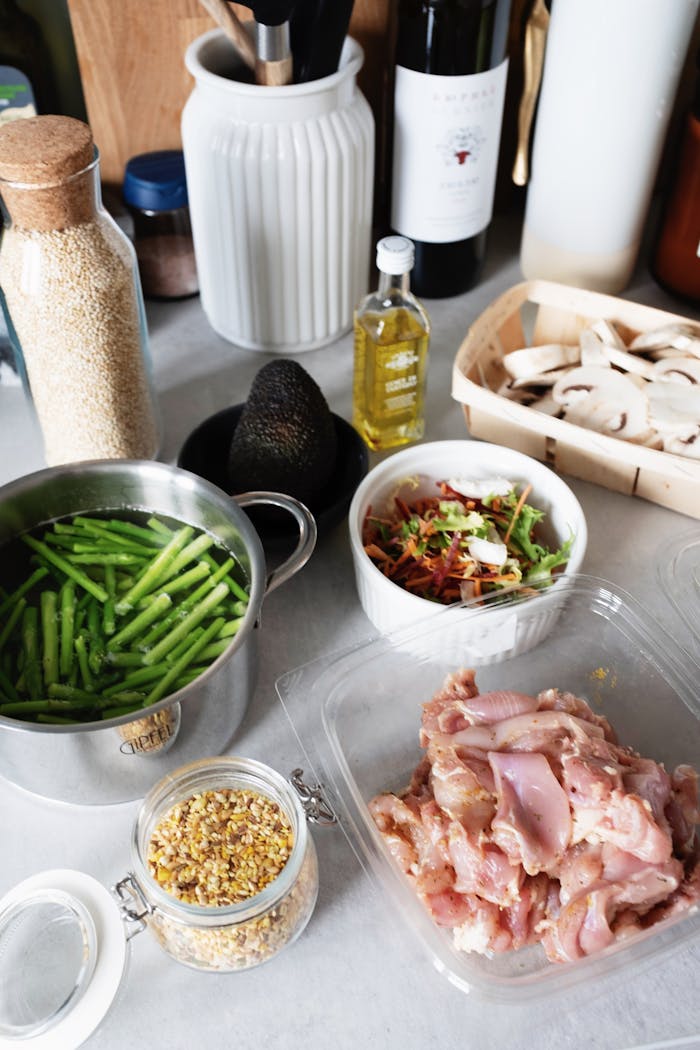 gallery-02 From above of raw chicken pieces in container green beans in pot of water and chopped champignons slices in wicker basket next to salad bowl avocado and spices in glass jar on countertop with various kitchen utensils